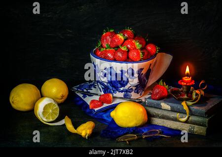 Stillleben mit leckeren frischen Erdbeeren in Porzellanschüssel mit frischen Zitronen, alten Büchern und beleuchteten Kerze auf dunklem Hintergrund platziert. Stockfoto
