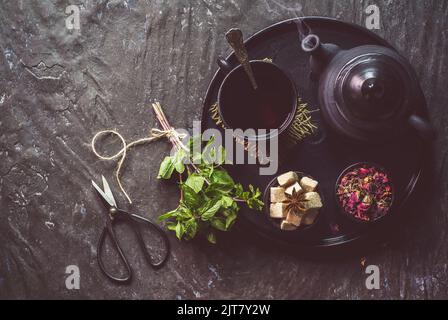 Heiße Tasse Tee platziert Tee mit Wasserkocher, Zucker, Minze und trockenen Blumen auf dunklem Hintergrund. Draufsicht mit Kopierbereich. Stockfoto