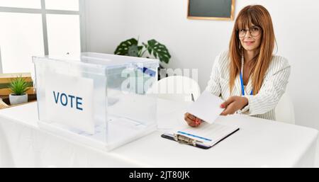 Junge hispanische Frau lächelt selbstbewusst und hält Stimmabgabe und schreibt in die Zwischenablage am Wahlkollegium Stockfoto