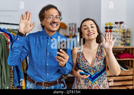 Interrassisches Paar im mittleren Alter im Einzelhandelsgeschäft, das Smartphone verwendet und dabei auf ein glückliches und lächelndes Hallo verzichtet, freundliche Willkommensgeste Stockfoto