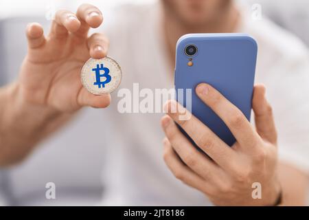 Junger kaukasischer Mann mit Smartphone hält Bitcoin zu Hause Stockfoto