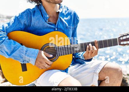 Mann, der klassische Gitarre spielt, sitzt am Strand auf einem Felsen. Stockfoto