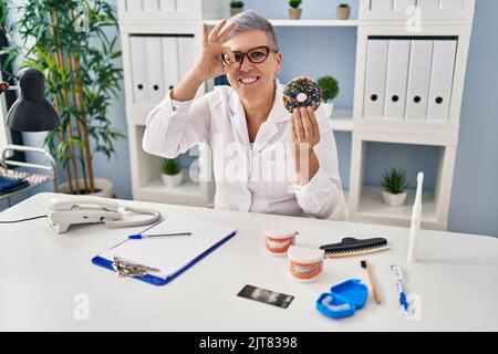 Mittlere Alter Zahnarzt Frau hält Schokolade Donut lächelnd in der Liebe tun Herz Symbol Form mit Händen. Romantisches Konzept. Stockfoto