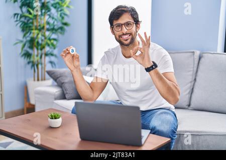 Schöner lateinischer Mann hält virtuelle Währung Bitcoin mit Laptop tun ok Zeichen mit den Fingern, lächelnd freundlich gestikulieren ausgezeichnetes Symbol Stockfoto