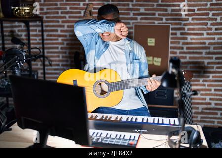 Hispanischer junger Mann, der im Musikstudio klassische Gitarre spielt, lächelt fröhlich und guckelt mit Händen, die Gesicht zeigen. Überrascht und begeistert Stockfoto