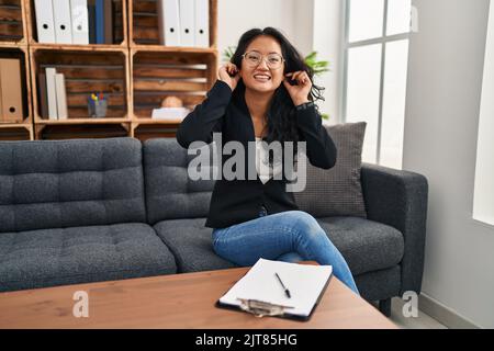 Junge asiatische Frau im Beratungsbüro lächelnd ziehen Ohren mit Fingern, lustige Geste. Vorsprechen Problem Stockfoto