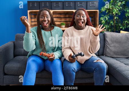 Zwei afrikanerin spielen Videospiel mit gehobener Hand und feiern den Sieg mit glücklichen Lächeln und dem Siegerausdruck Stockfoto