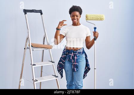 afroamerikanische Frau hält Roller Maler lächelnd und zuversichtlich gestikulieren mit der Hand tun kleine Größe Zeichen mit Fingern suchen und die Kamera. mea Stockfoto