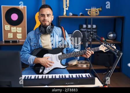 Junger hispanischer Musiker, der mit einem Lächeln auf den Lippen lächelt und im Musikstudio elektrische Gitarre spielt Stockfoto