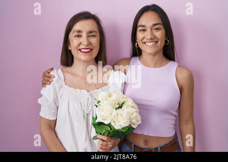 Hispanische Mutter und Tochter halten Strauß von weißen Blumen mit einem glücklichen und kühlen Lächeln auf dem Gesicht. Glückliche Person. Stockfoto