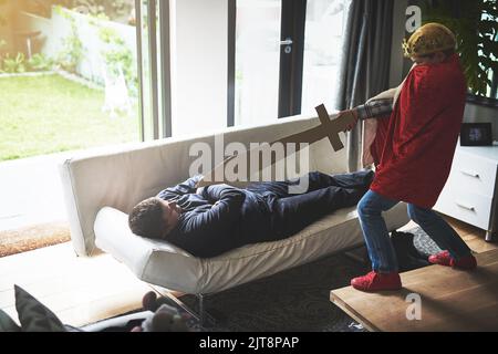 Der Sieg gehört mir. Ein kleiner Junge in einem königlichen Kostüm, der vorgibt, seinen schlafenden Vater mit einem Schwert anzugreifen. Stockfoto