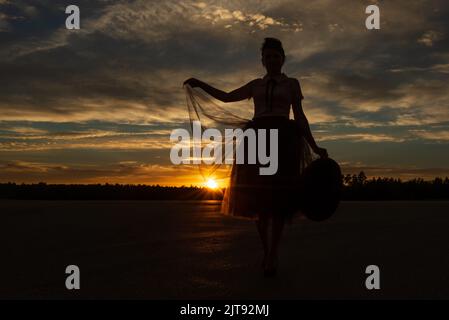 Low-Winkel-Ansicht der Märchenfrau halten Kante des Tutu Rock durch die Sonne gegen Wald Silhouette und Sonnenuntergang bewölkten Himmel mit Hut in der Hand hinterleuchtet Stockfoto