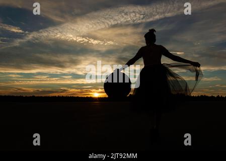 Silhouette einer märchenhaften Frau, die gegen den Sonnenuntergang steht, mit ausgestreckten Armen, die ihren Hut und den Rand des Tutu-Rockes gegen den Wald und den wolkigen Himmel hält Stockfoto