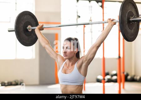 Muskulöse Sportlerin mit Pferdeschwanz, der wegschaut und mit schwerer Langhantel während des intensiven Gewichtheben-Workouts auf verschwommenem Rücken eine Schnatchübung macht Stockfoto