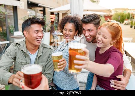 Eine Gruppe von glücklichen und vielfältigen Freunden klirren mit Bier, während sie gemeinsam auf der Terrasse des Cafés im Freien in der Stadt Zeit verbringen Stockfoto