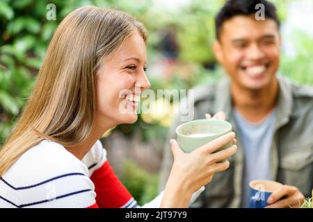 Positive Frau mit einer Tasse Cappuccino auf der Terrasse in der Nähe eines asiatischen Mannes auf verschwommenem Hintergrund beim Frühstück am Sommermorgen Stockfoto