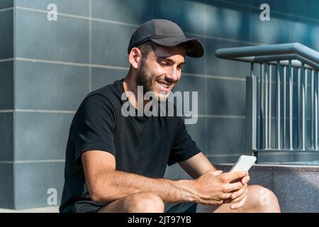 Fröhlicher Mann in legerer Kleidung und Mütze lächelnd und SMS-Nachrichten in den sozialen Medien auf dem Handy zu senden, während er auf Treppen vor dem modernen Gebäude sitzt Stockfoto