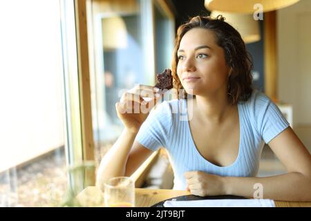 Eine Frau, die einen Schokoladenkuchen hält und isst, sitzt in einem Restaurant und schaut durch ein Fenster Stockfoto