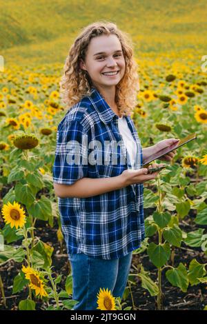 Porträt eines lächelnden lockigen Mädchens, das im Sonnenblumenfeld arbeitet und die Analyse des Wachstums der Pflanzenkultur macht. Intelligente Landwirtschaft und Präzisionslandwirtschaft Stockfoto