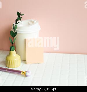 Papierkaffeetasse, Notizblock, Stift, trockene Pflanze auf weißem Schreibtisch. Rosa Wandhintergrund Stockfoto