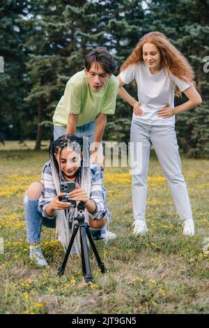 Eine Gruppe von drei jungen Teenagern, die ein Gruppen-Selfie mit einem Selfie-Stick machen wollen und im Stadtpark stehen. Social-Media-Trends. Freundschaftstag. Stockfoto