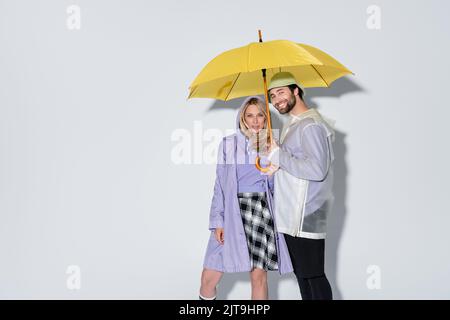 Frau in Tartan Rock stehend mit glücklichen Mann in panama Hut unter gelben Regenschirm auf grau, Stock Bild Stockfoto