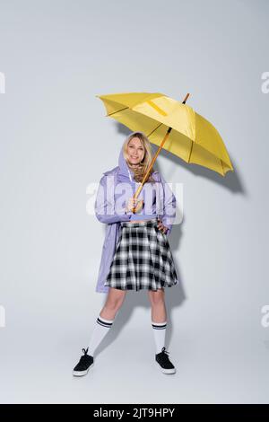 Volle Länge der glücklichen Frau in Tartan Rock und Longs Socken mit Sneakers stehen unter Regenschirm auf grau, Stock Bild Stockfoto