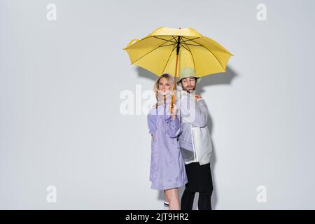 Blonde Frau in Tartan Rock stehend mit bärtigen Mann in panama-Hut unter gelben Regenschirm auf grau, Stock Bild Stockfoto