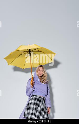 Glückliche Frau in schartanem Rock lächelnd stehend unter gelbem Regenschirm auf grauem, Stockbild Stockfoto