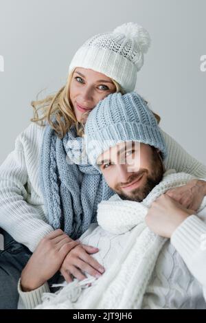 Fröhliche Frau in gestrickten Schal umarmt bärtigen Freund im Winter Hut isoliert auf grau, Stock Bild Stockfoto