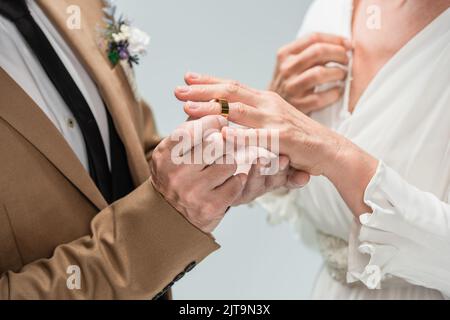 Beschnittene Ansicht des Bräutigams mit goldenem Verlobungsring am Finger der Braut in weißem Kleid isoliert auf grau, Stockbild Stockfoto