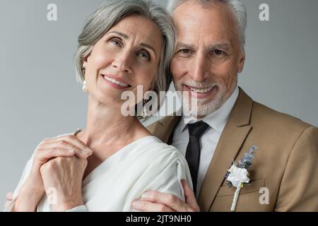 Portrait von glücklichen Mann mittleren Alters in Anzug mit floralen boutonniere umarmende Frau in weißem Hochzeitskleid isoliert auf grau, Stockbild Stockfoto
