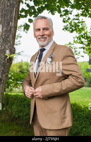 Bärtiger Bräutigam mittleren Alters, beigefarbener Blazer mit Boutonniere und Lächeln im grünen Park, Stockbild Stockfoto