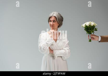 Bräutigam hält Hochzeit Strauß in der Nähe verwirrt mittleren Alters Braut in weißem Kleid isoliert auf grau, Stockbild Stockfoto