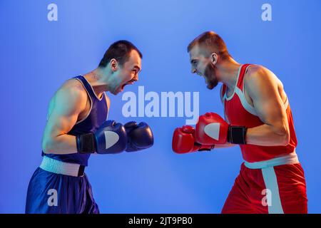 Sportliche Emotionen. Aufgeregt professionelle Boxer in rot und blau Sport Uniform Boxen isoliert auf blauem Hintergrund in Neonlicht. Sport, Geschicklichkeit, Kraft Stockfoto