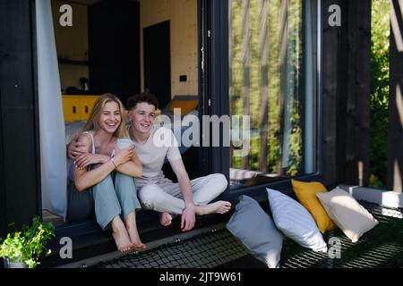 Junges Paar sitzt mit einer Tasse Kaffee und kuschelt auf der Terrasse in ihrem neuen Zuhause in einem winzigen Haus im Wald, nachhaltiges Wohnkonzept. Stockfoto