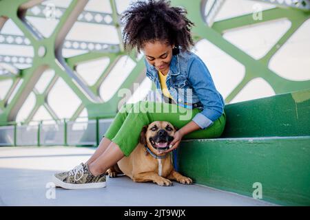 Multirassisches Mädchen, das mit ihrem Hund draußen auf der Brücke sitzt und sich ausruht, ihn trainiert und gemeinsam Freizeit verbringt. Konzept der Beziehung zwischen Stockfoto