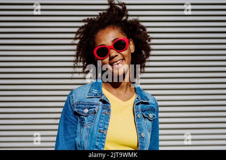 Porträt eines jungen glücklichen multirassischen Teenagers mit Sonnenbrille und Afro-Frisur, im Freien stehend. Stockfoto