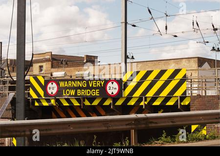 Bild der sehr niedrigen Straßenbrücke, 2,7m, neben dem Ely Bahnhof, Ely Cambridgeshire mit einer Armco-Crashbarriere und elektrischen Zugdrähten Stockfoto