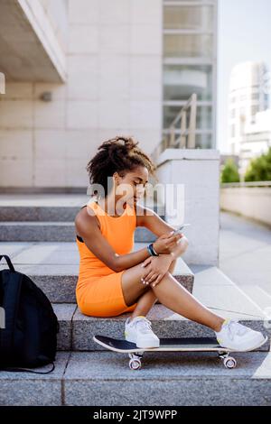 Multirassisches Teenager-Mädchen mit Rucksack und Skateboard, sitzend und mit Smartphone in der Stadt während des Sommers. Stockfoto