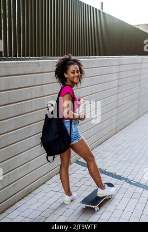 Multirassisches Teenager-Mädchen mit Rucksack am Skateboard, während des Sommers. Stockfoto