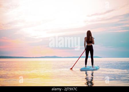 Rückansicht eines Surfers mit Mädchen, das bei Sonnenaufgang auf dem Surfbrett auf dem See paddelt. Stockfoto