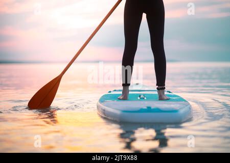 Rückansicht eines Surfermädchen, das bei Sonnenaufgang auf dem Surfbrett auf dem See paddelt, unterer Abschnitt. Stockfoto