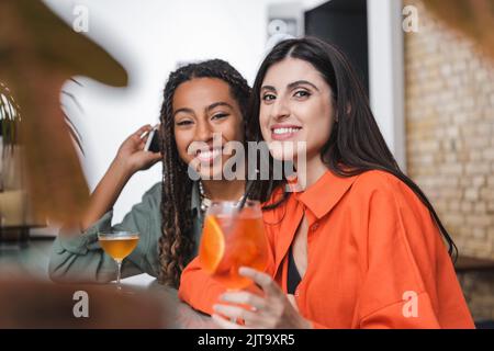 Lächelnde multiethnische Freundinnen mit Smartphone, das in der Nähe von Cocktails im Café auf die Kamera schaut, Stockbild Stockfoto