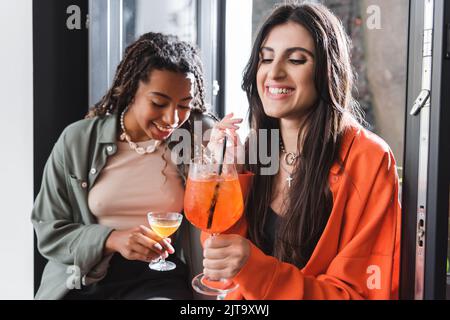 Lächelnde Frau mit Cocktail in der Nähe der verschwommenen afroamerikanischen Freundin und Fenster im Café, Stockbild Stockfoto