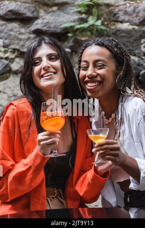 Lächelnde afroamerikanische Frau mit Cocktail in der Nähe der Freundin auf der Terrasse des Cafés, Stockbild Stockfoto