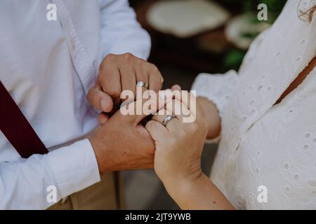 Nahaufnahme von Braut und Bräutigam, die beim Empfang draußen im Hinterhof Hände mit Eheringen halten. Stockfoto