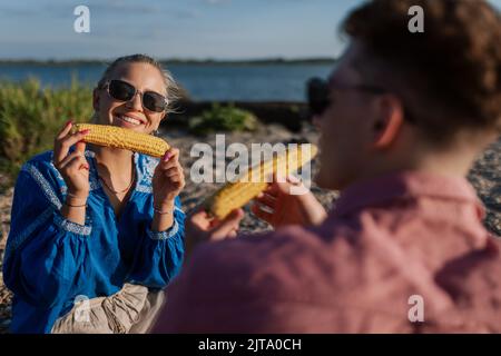 Glückliches junges Paar, das am Strand zusammen auf einer Decke sitzt und Mais isst. Genießen Sie die gemeinsame Urlaubszeit. Stockfoto