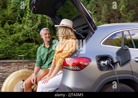 Ein Paar mittleren Alters sitzt im Kofferraum, während es auf das Aufladen des Autos wartet, bevor es in den Sommerferien reist. Stockfoto