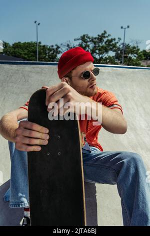 Junger und trendiger Mann in Sonnenbrille sitzt mit Skateboard und schaut weg Stockfoto